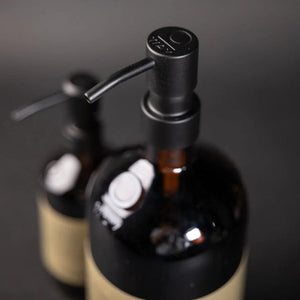 Two Oway Reusable Metal Pump Dispensers from Simply Colour Hair Salon Studio & Online Store are pictured—one sharply in the foreground, the other blurred behind—highlighting their sleek and reusable brown bottle design against a dark backdrop.