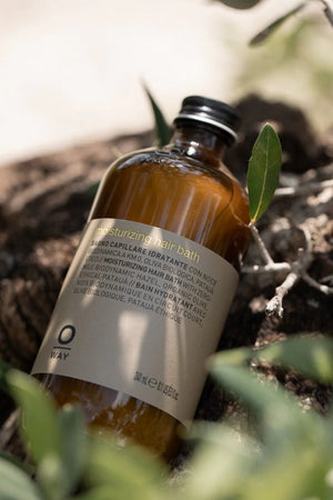 A brown Oway Moisturizing Hair Bath bottle labeled "Hydrating Shampoo for Dry, Lackluster Hair" sits on a natural surface outdoors, partly framed by green leaves. The multilingual label highlights its moisturizing benefits.
