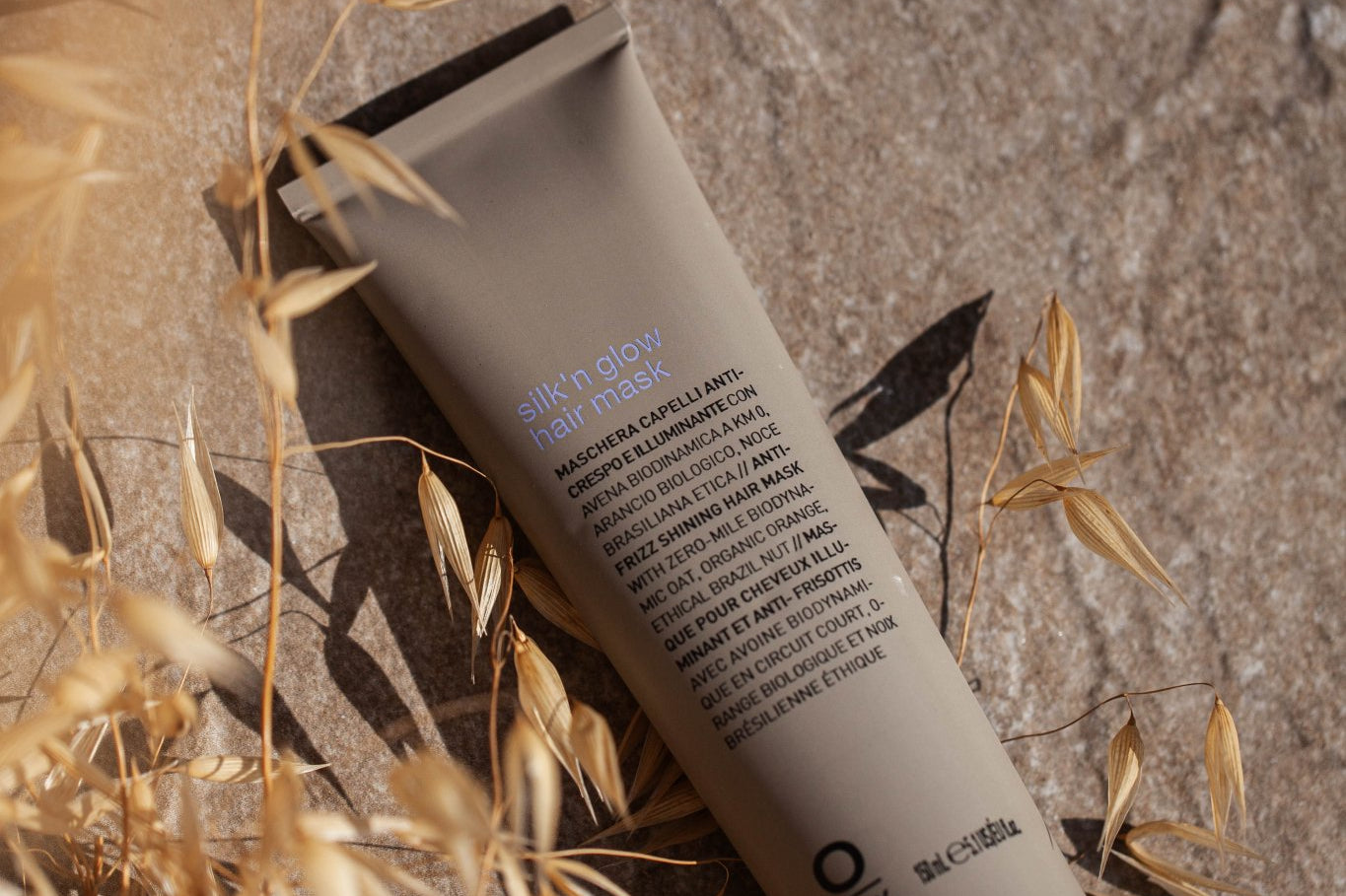 A tube of Oway Silk’N Glow Hair Mask | Nourishing Hair Mask for Dry, Frizzy Hair lies on a stone surface with dried grass and sunlight, ideal for taming dry, frizzy hair with organic Cupuaçu butter.