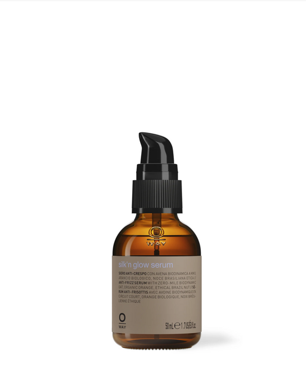 A brown glass bottle with a black pump labeled “Oway Silk’N Glow Serum | Anti-Frizz Shine Serum for Dry, Frizzy Hair” holds 50 ml (1.69 fl oz) and features product information and ingredients on the label.