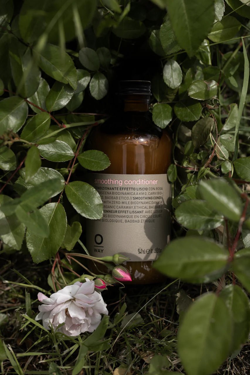 A brown bottle of Oway Smoothing Conditioner | Detangling Conditioner for Soft, Polished Hair sits amid green leaves and grass with a pale pink flower—ideal for taming frizz and enriched with nourishing baobab oil.