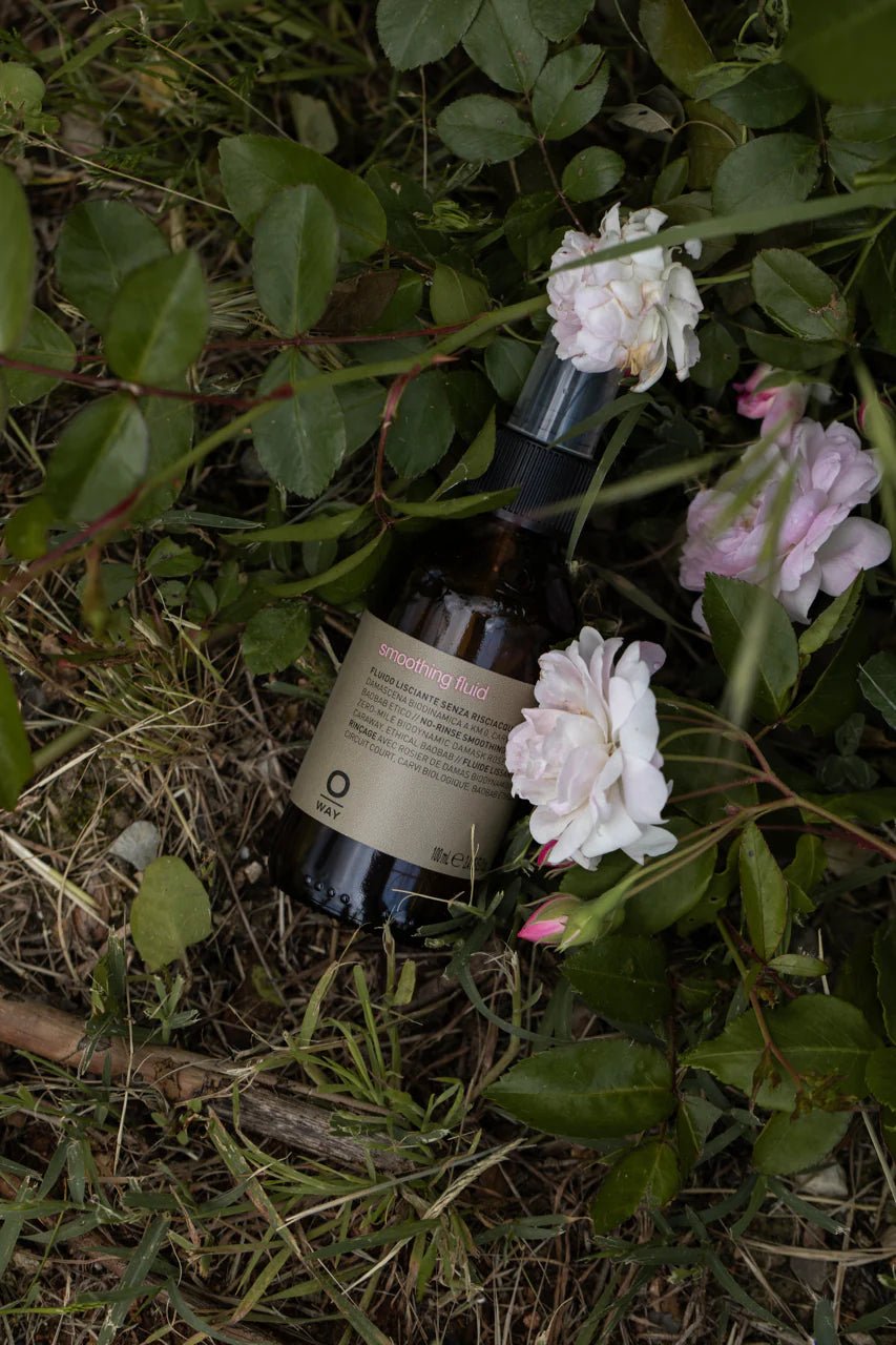 A bottle of Oway Smoothing Fluid | Sleek Styling Serum for Smooth, Frizz-Free Hair rests on grass and soil among green leaves and pale pink roses, promising shiny, sleek hair.