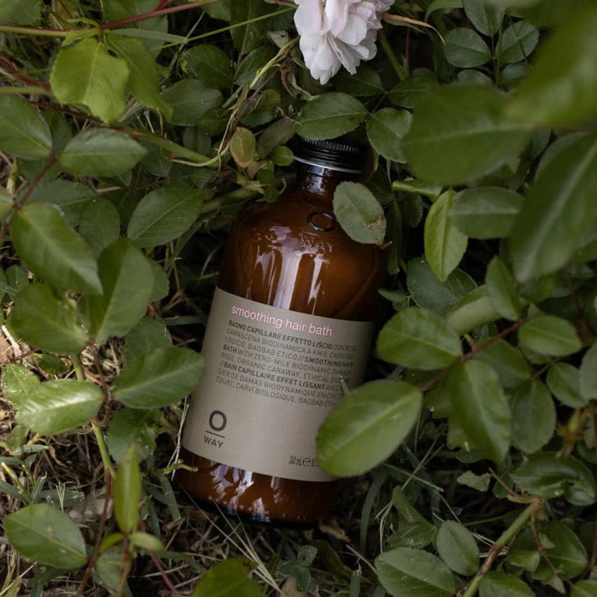 A brown bottle of Oway Smoothing Hair Bath, a frizz-control shampoo for coarse or unruly hair, is nestled among green leaves and an organic rose in a garden setting, suggesting gentle smoothing by Oway.