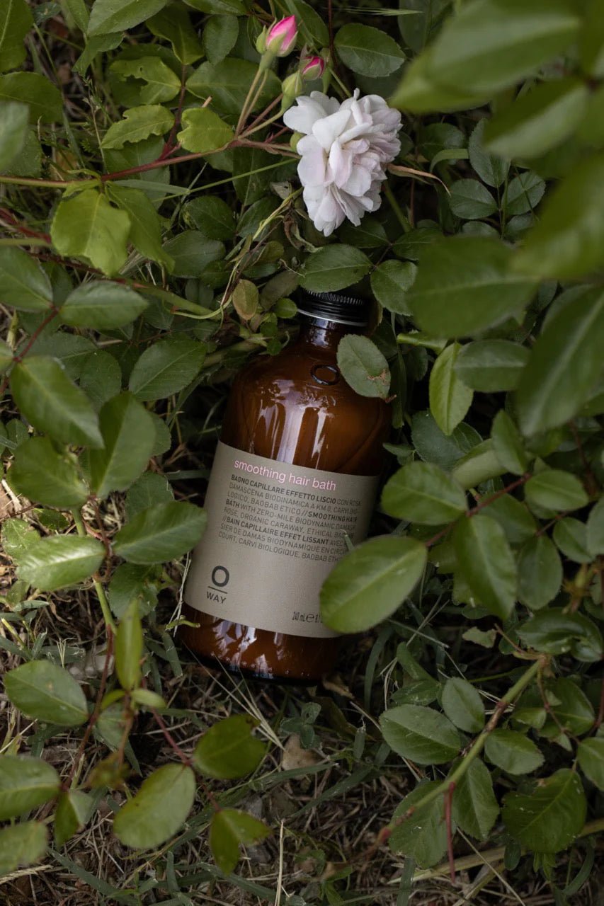 A brown bottle of Oway Smoothing Hair Bath, a frizz-control shampoo for coarse or unruly hair, is nestled among green leaves and an organic rose in a garden setting, suggesting gentle smoothing by Oway.