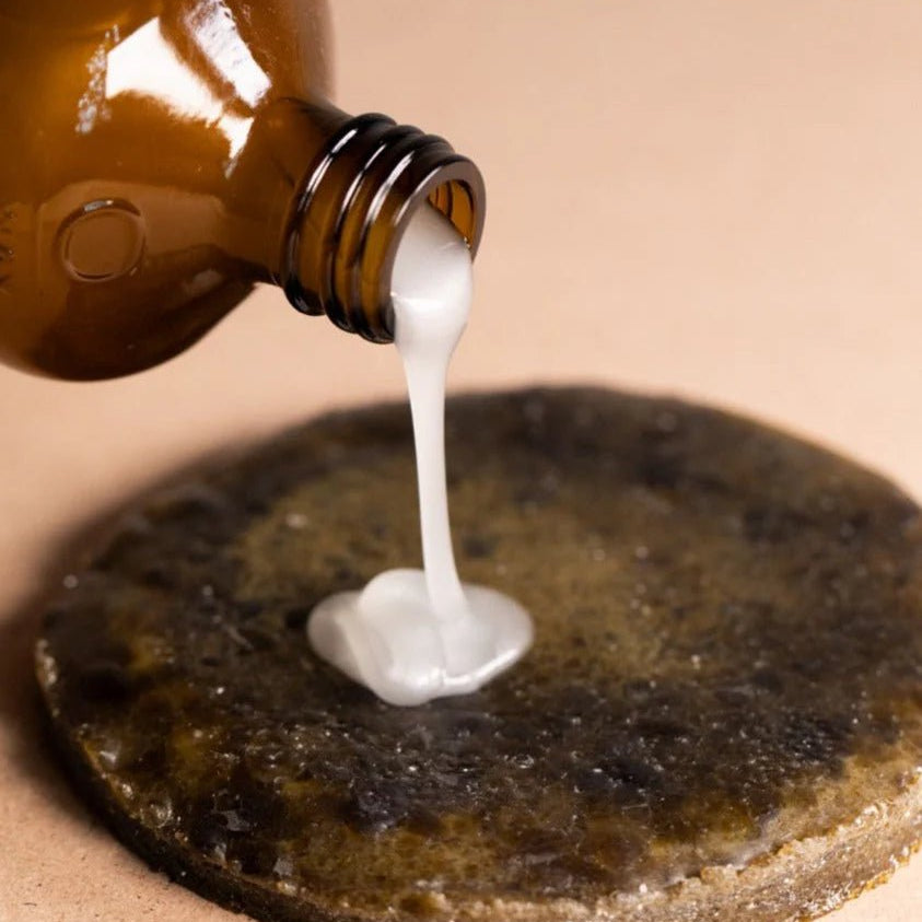 A brown Oway bottle pours Smoothing Hair Bath—an anti-frizz shampoo for coarse or unruly hair—onto a dark textured surface with a tan backdrop, perfect for smoothing and taming frizzy hair.