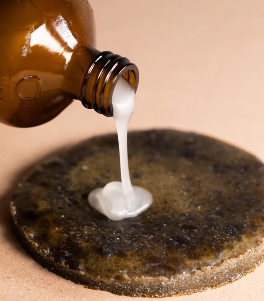 A brown Oway bottle pours Smoothing Hair Bath—an anti-frizz shampoo for coarse or unruly hair—onto a dark textured surface with a tan backdrop, perfect for smoothing and taming frizzy hair.