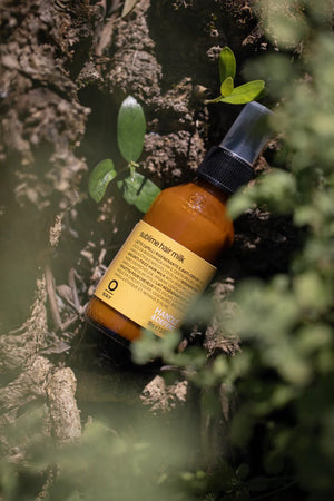 A brown bottle of Oway Sublime Hair Milk | Lightweight Leave-In Moisture Milk for Softness and Shine with a pump top stands among green plants and tree bark, photographed outdoors in natural light.