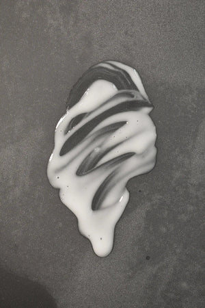 A small puddle of white liquid with streaks sits on a smooth, gray surface—resembling Oway Sublime Hair Milk, a lightweight leave-in moisture milk by Oway that delivers softness and shine while controlling frizz.