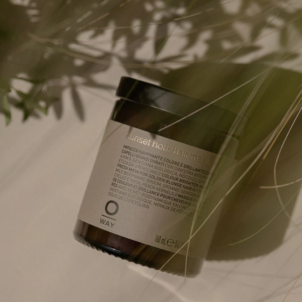 A jar of Oway Sunset Hour Hair Mask | Radiance-Boosting Mask for Golden Blonde Hair sits on a neutral surface, partially shaded by foliage.