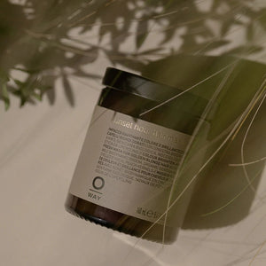 The Oway Sunset Hour Hair Mask, a radiance-boosting mask for golden blonde hair, sits on a light surface with shadows and green leaves partially obscuring the view. The jar highlights its color-enhancing formula with biodynamic broom.