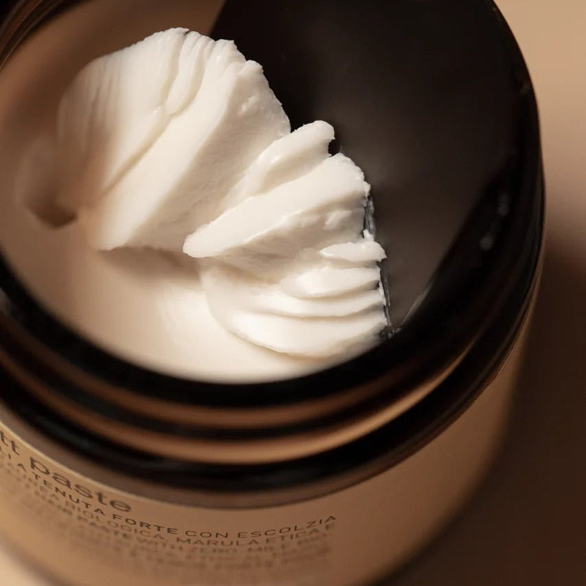 A close-up of Oway Supreme Matte Paste, a strong hold styling paste with a matte finish, as a black spatula scoops some of the product from its brown jar.
