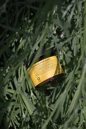 A jar of Oway Supreme Matte Paste | Strong Hold Styling Paste with Matte Finish, featuring a yellow label, lies on its side among tall green grass, partly shaded by the blades.