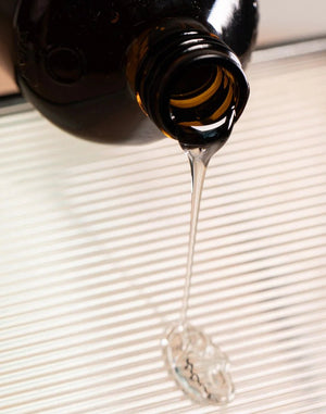 A close-up of Oway Volumizing Hair Bath, a gentle volumizing shampoo for fine hair, being poured from a dark brown glass bottle onto a white ridged surface. The organic ginger-infused liquid forms a small pool below.