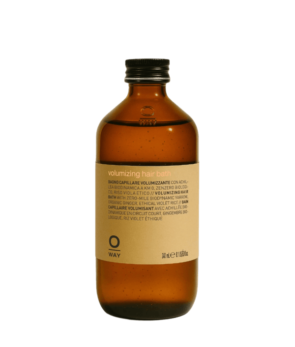A brown glass bottle with a black cap and a beige "Oway Volumizing Hair Bath" label for fine or limp hair, featuring multilingual product info, sits against a green textured background. Brand: Oway.