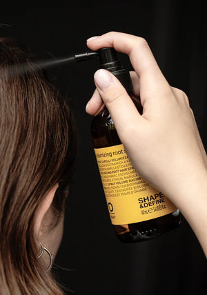 A person sprays Oway Volumizing Root Spray | Lightweight Root Lift for Fine, Flat Hair onto their hair, holding the brown bottle with their head turned away from the camera against a plain black background.