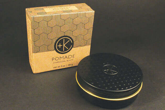 A Cult and King POMADE CHARCOAL BLACK | Black Pepper & Lavender Clay-Paste Hybrid container with a gold lid sitting on a table, hold and separation.