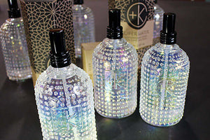 Three iridescent, studded glass bottles with black caps display Cult and King SUPER WATER, an antioxidant toner for hair and skin. Elegant packaging in the background evokes vegan luxury by Cult and King.