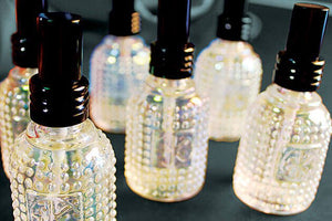 Six clear glass bottles with textured surfaces and black spray tops are shown on a dark surface. Each contains Cult and King SUPER WATER, a vegan antioxidant toner for hair and skin to boost hydration, displayed in close-up detail.
