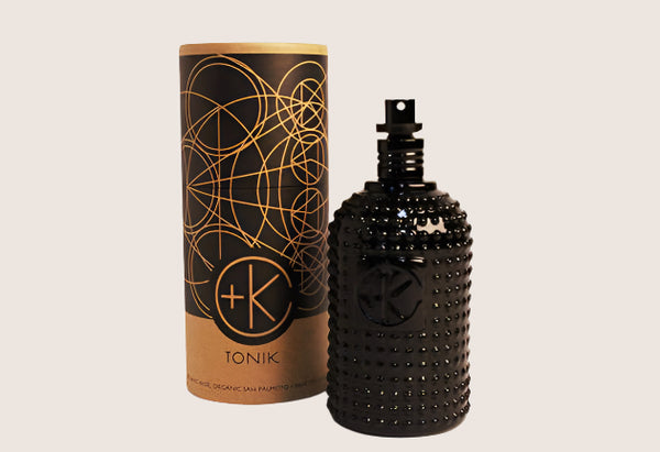 A black textured bottle of Cult and King TONIK | Conditioning Hair & Scalp Potion with a spray nozzle stands before a cylindrical container with gold geometric patterns and the text "+K TONIK" against an off-white background.