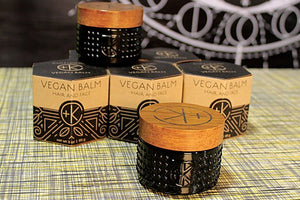 Five Cult and King VEGAN BALM | Manifest Moisture containers for hair styling and face are displayed on a checkered surface—three in branded boxes, two unboxed wooden-lidded jars—with a blurred background enhancing focus.