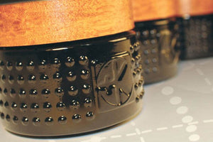 Close-up of Cult and King VEGAN BALM, a cylindrical hair styling tool with a textured black base featuring raised dots and an embossed Cult and King logo, plus a smooth wood-toned top. It rests on a faintly grid-patterned surface.