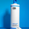 A large white bottle of K18 Hair Repair K18 Damage Shield pH Protective Conditioner with a blue pump on a blue background, next to a dollop of conditioner, emphasizing hair health and damage protection.
