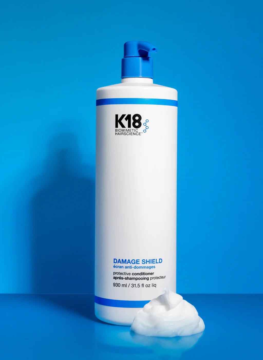 A 930 ml bottle of K18 Hair Repair K18 Damage Shield pH Protective Conditioner - Multiple Sizes, enriched with K18PEPTIDE™, stands beside a dollop of the product against a blue background, promoting hair health.