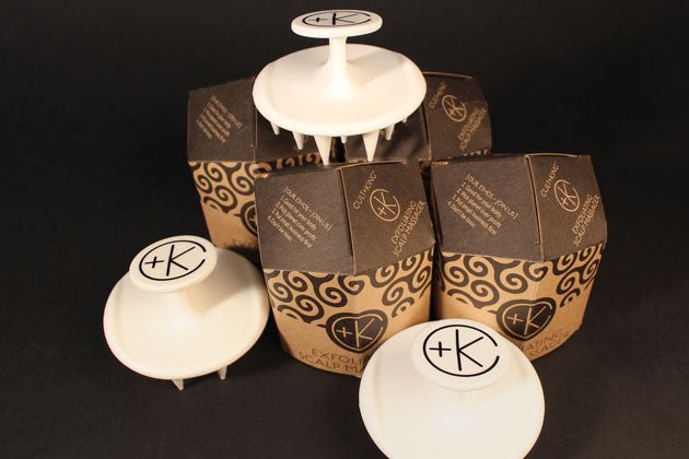Several boxed Cult and King Exfoliating Scalp Massagers are displayed against a black background. Three massagers are out of their boxes, revealing their white color and spiked design. These silicone scalp brushes from Cult and King feature firm yet flexible bristles for effective exfoliation of the scalp.