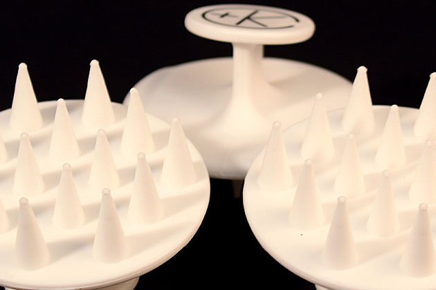 Three white Cult and King Exfoliating Scalp Massagers, circular plastic objects with pointed spikes, are shown against a dark background; one of them features a handle.