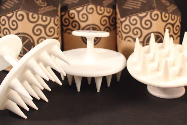 Three Cult and King Exfoliating Scalp Massagers, made of white plastic and featuring firm yet flexible bristles, are displayed against a background of patterned brown packaging.