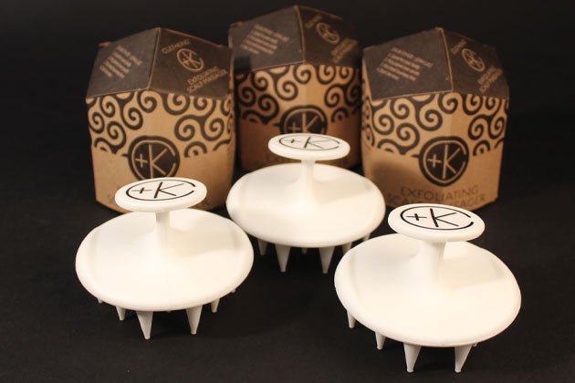 Three Cult and King Exfoliating Scalp Massagers with handles, each featuring the Cult and King logo, are displayed in front of three product packaging boxes with a swirling design. These white silicone scalp brushes boast firm yet flexible bristles for an invigorating experience.