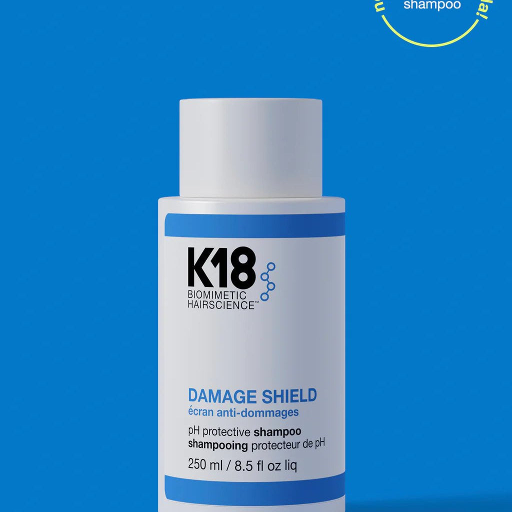 A 250 ml bottle of K18 DAMAGE SHIELD pH Protective Shampoo - Multiple Sizes by K18 Hair Repair is displayed against a blue background. A circular label at the top right reads "new name! same formula! pH maintenance shampoo," featuring K18PEPTIDE™ for daily use.