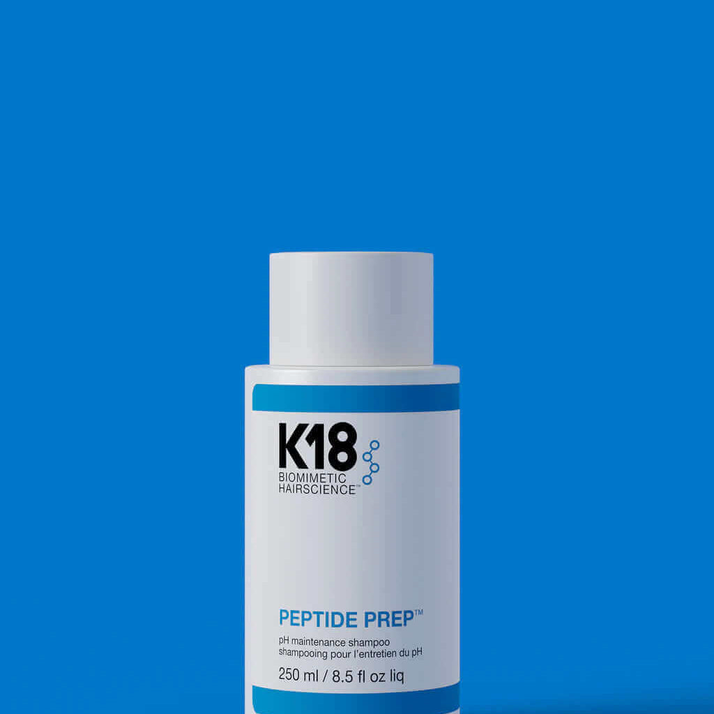 A white bottle labeled "K18 DAMAGE SHIELD pH Protective Shampoo - Multiple Sizes" stands against a blue background. This 250 ml (8.5 fl oz) cleansing shampoo, crafted with K18PEPTIDE™, is perfect for daily use to maintain optimal pH balance and hair health.