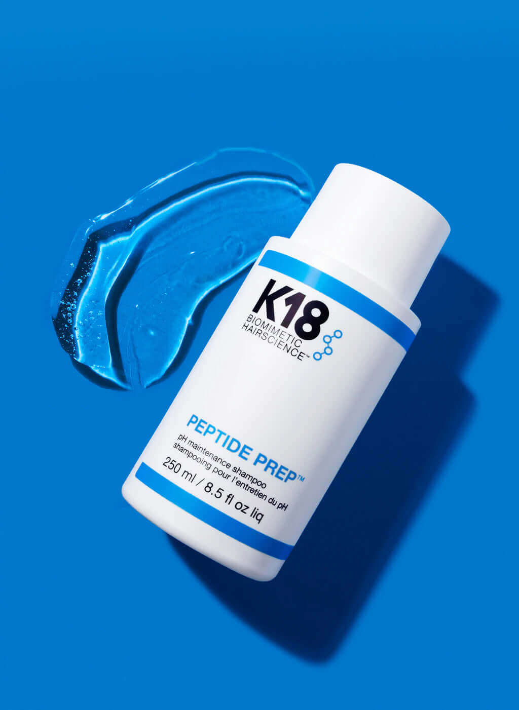 White bottle of K18 DAMAGE SHIELD pH Protective Shampoo - Multiple Sizes by K18 Hair Repair on a blue background, with a smear of the gel-like product beside it. Suitable for daily use to revitalize your hair, powered by the K18PEPTIDE™ formula.