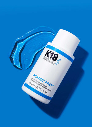 A white bottle of K18 Hair Repair DAMAGE SHIELD pH Protective Shampoo - Multiple Sizes rests on a vibrant blue background beside a smear of blue gel, emphasizing its pH-balanced formula.