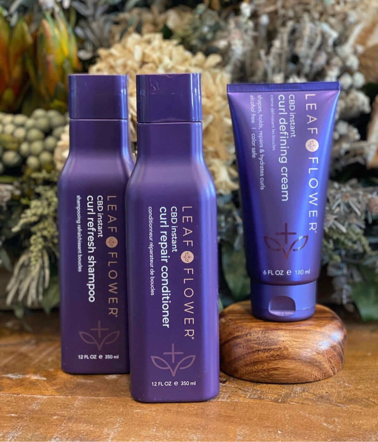 Three bottles of Leaf and Flower Instant Curl Defining Cream, specializing in frizz reduction and hydration, are sitting on a wooden table.