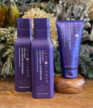Three purple bottles of Leaf and Flower hair products—Curl Refresh Shampoo, Curl Repair Conditioner, and Instant Curl Defining Cream—stand upright on a wooden surface, highlighting frizz-free curl solutions with a floral arrangement behind them.