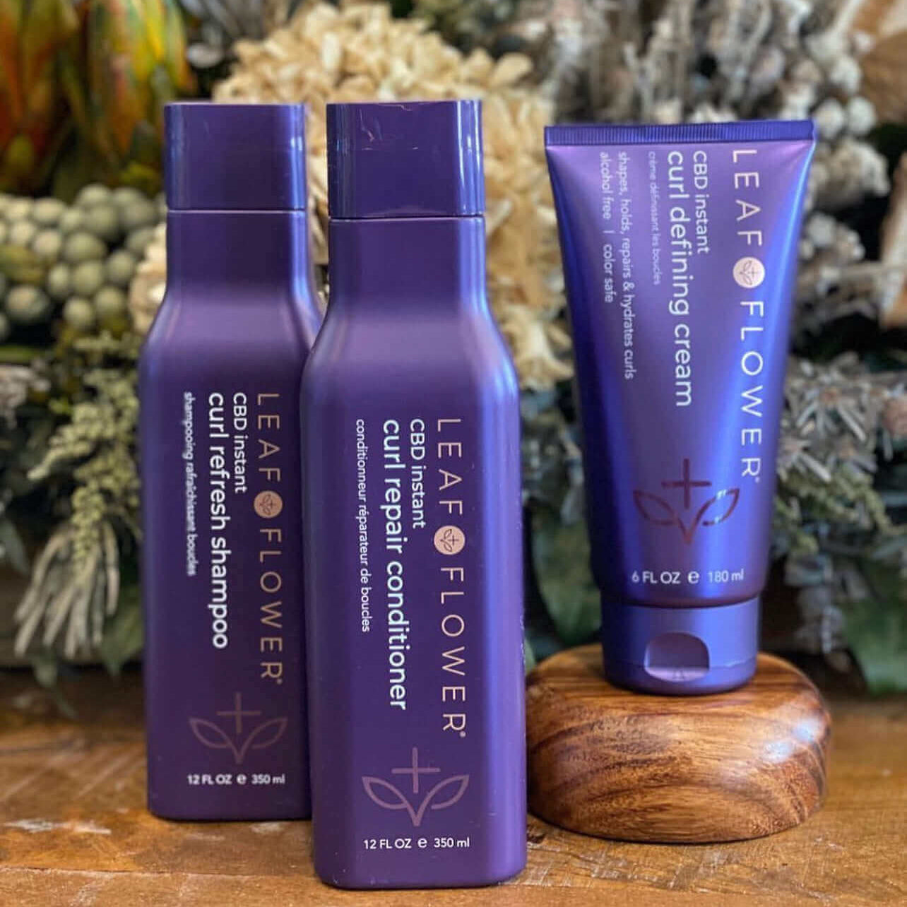 Three purple Leaf and Flower hair care products—Instant Curl Repair Conditioner (multiple sizes), curl refresh shampoo, and curl defining cream—are elegantly arranged on a wooden surface with lush plants in the background.
